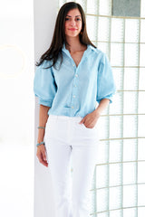 A model wearing the Finley Bomba blouse, a light teal  button-down shirt with elbow blouson sleeves and a front twist hem detail.