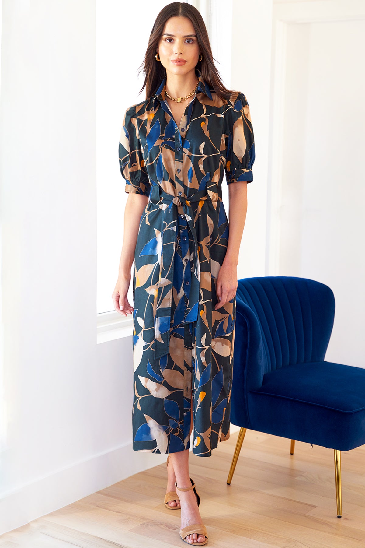 A model wearing the Harper shirt dress, featuring a gold and navy floral print, tie-front, pockets, and made from 100% cotton with a point collar.
